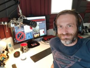 Dirk Primbs mit Kopfhörern an seinem Schreibtisch, daneben ein Mikrofon am Gelenkarm und ein Monitor mit geöffneter Audiosoftware.