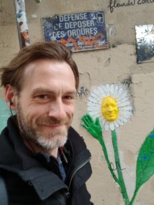 Dirk Primbs als Selfie vor einer Hauswand mit aufgemalter Margerite und einem französischen Verbotsschild.