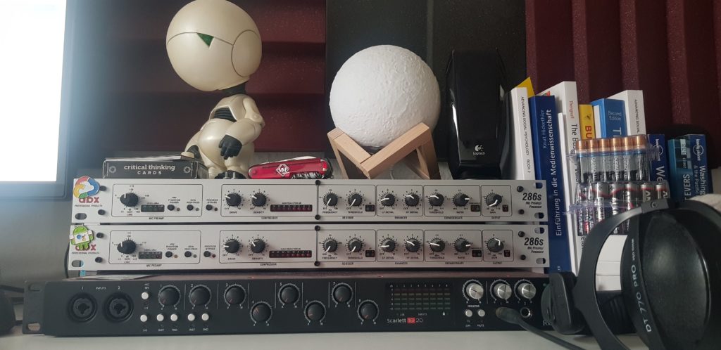 Zwei übereinander gestapelte dbx-286s-Kanalzüge über einem Focusrite-Scarlett-18i20-Audiointerface, daneben Bücher, eine Mondlampe, eine Marvin-Figur und Beyerdynamic-Kopfhörer.