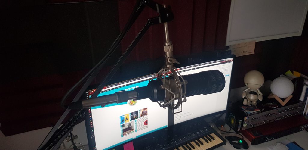 Ein großes dynamisches Mikrofon in einer Spinne am Gelenkarm vor einem Monitor mit geöffneter Audiosoftware, darunter ein MIDI-Keyboard; an den Wänden Akustikelemente.