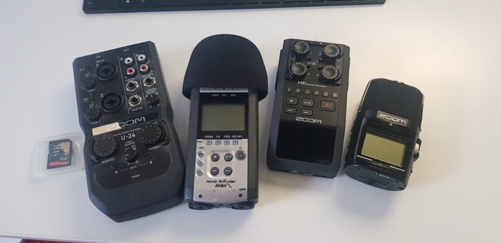 Vier Zoom-Geräte nebeneinander auf einem Tisch: ein U-24-Audiointerface, ein H4n mit aufgestecktem Windschutz, ein H6 und ein kleinerer Handrekorder, daneben eine SD-Karte im Etui.