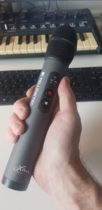 Eine Hand hält ein schwarzes Handmikrofon mit Sennheiser-Schriftzug; im Hintergrund ein Keyboard und eine Computertastatur.